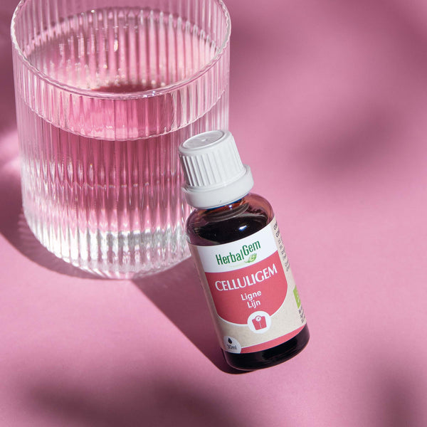 Flacon Celluligem Bio HerbalGem pour la ligne, de 30 ml, posé à côté d'un verre d'eau, sur fond rose.