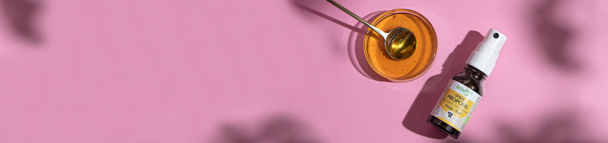 Collection propolis HerbalGem, spray propolis bio avec bol de miel et cuillère sur fond rose.